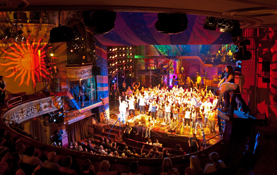 Hair at Gielgud Theatre. Photo Credit: Ian Grundy & Theatres Trust Taken from the theatre balcony, you see an aerial view of the audience watching a performance onstage with a large group of people dancing and singing.