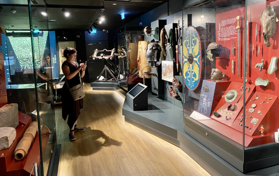 Trimontium Museum (Credit: Trimontium Trust) A woman looks at a display of old roman artefacts in a museum - including armour, shields and swords.