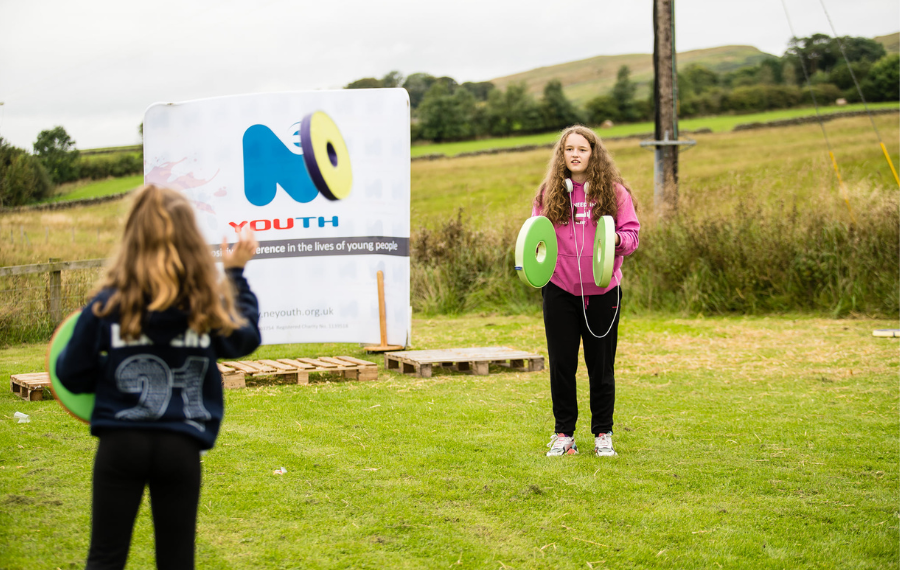 Young women playing sports outdoors – (Credit: NE Youth) Two young women stand a few metres apart and facing each outdoors in a field. They are playing a game where they throw large plastic discs back and forth on a lawn of grass in the sun.