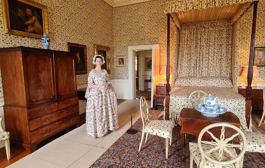 Principal Bedroom – Paxton House Trust A white woman in a old-style, flowery white dress stands in a 1770s bedroom with historic brown furniture, four poster bed and white flowery wallpaper.