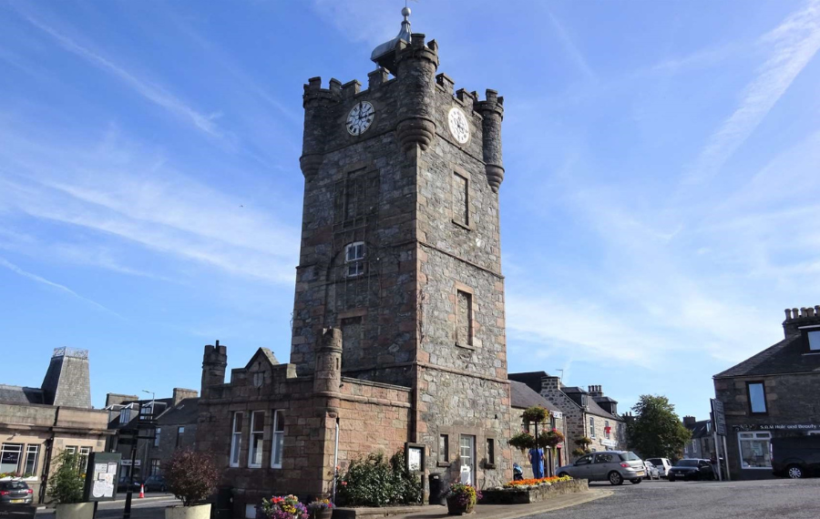 Dufftown Clocktower (c) Dufftown and District Community Association In the middle of a outdoor town square is a square tower, four storeys high, made of grey brick with a clock face at the top and turrets.
