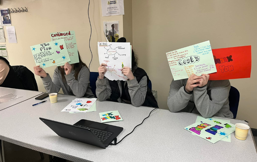 Young women’s workshop – (Credit: NE Youth) Three young women sit by a table and use the posters they've created to cover their faces and show what they've done. The posts read 'costs', 'plans' and 'ideas' and are mind maps of life planning.