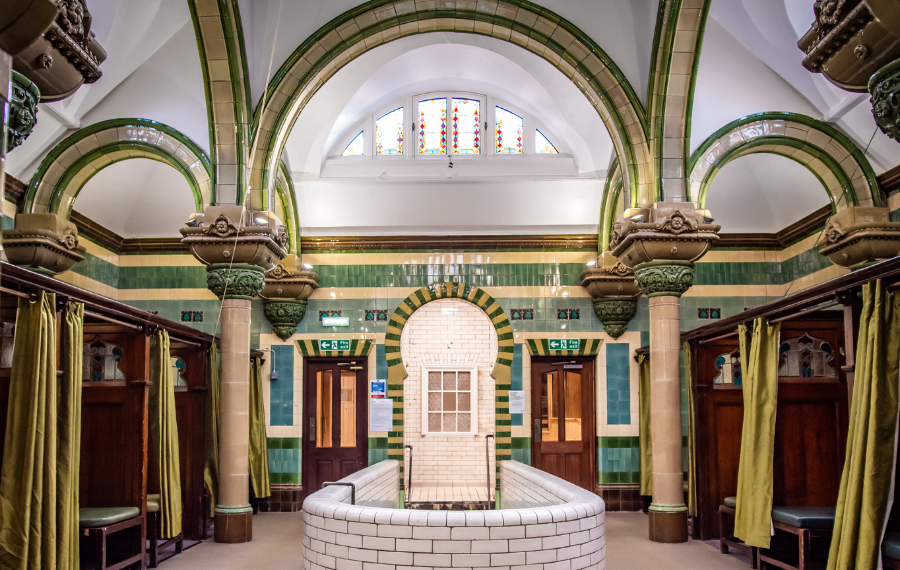 Carlisle Public Baths – Credit: Architectural Heritage Fund An old bathhouse with green, light green and white tiles, arches and columns, a white bath area and dark wood changing booths