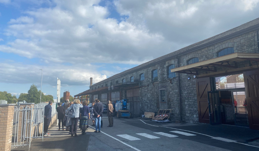 Llanelli Railway Goods Shed – Trustee Visit A group of adults of different ages stand into front of a one storey, long grey building listening to a guide speak.
