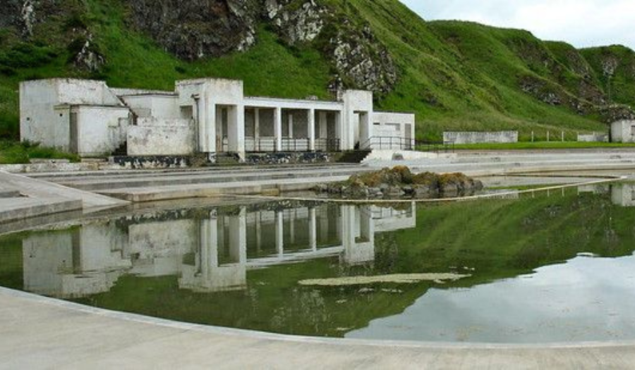 Tarlair Lido – (Credit: CC by NC 2.0) Panoramic view of Tarlair swimming pool. It's an outdoor lido with white block outhouses and beautiful green hills around it.
