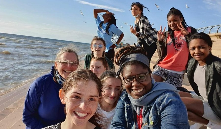 Day trip – (Credit: M13) Group of young girls pose for a selfie by the sea on a sunny day.
