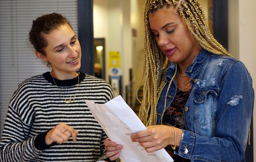 Young women working – (Credit: Odd Arts) Two young women stand in an office. One is showing the other writing on a piece of paper and the other is looking and talking about it.